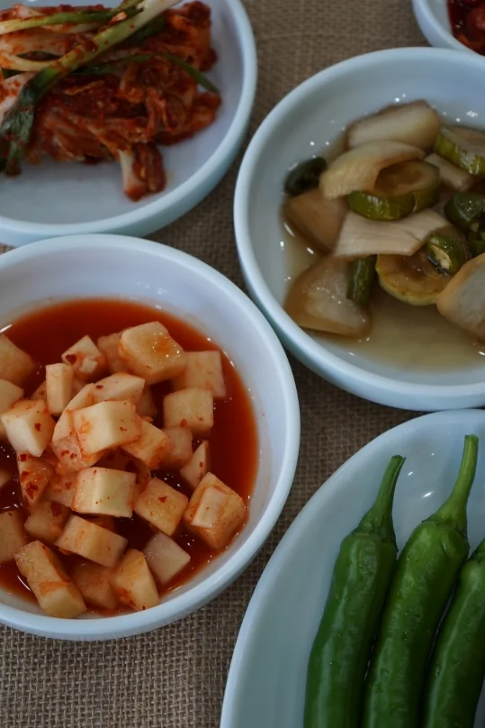 밑반찬 세로 컷 — 앞의 깍두기, 뒤로 김치·장아찌, 오른쪽 아래 풋고추