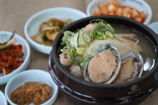 전복삼계탕 — 닭 위에 껍데기째 올린 전복과 파, 곁의 김치·장아찌·깍두기와 장