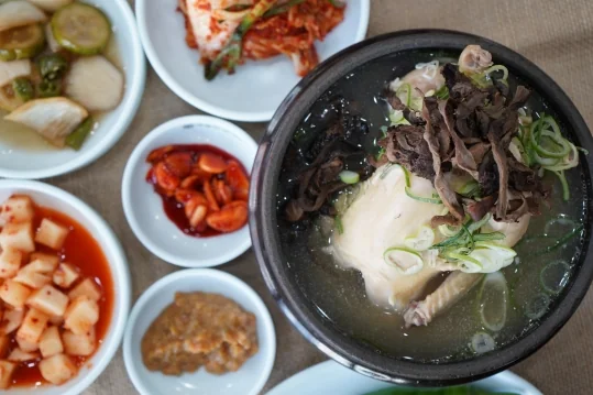 능이버섯삼계탕 한 상 — 위에서 본 뚝배기(닭 위 능이버섯)와 김치·장아찌·깍두기·붉은 양념 반찬·장