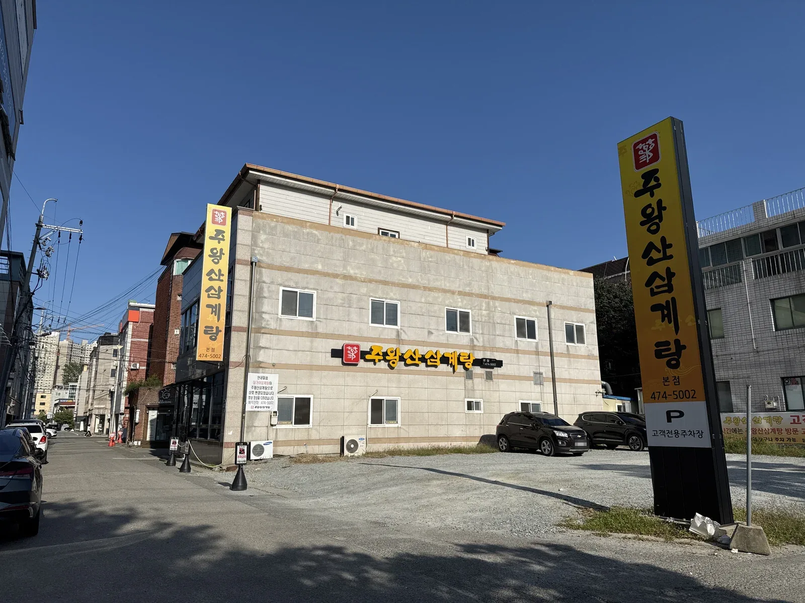 식당 건물과 자갈 주차장 — 벽의 노란 간판 «주왕산삼계탕», 오른쪽 기둥 간판 «주왕산삼계탕 본점 474-5002 · P 고객전용주차장»
